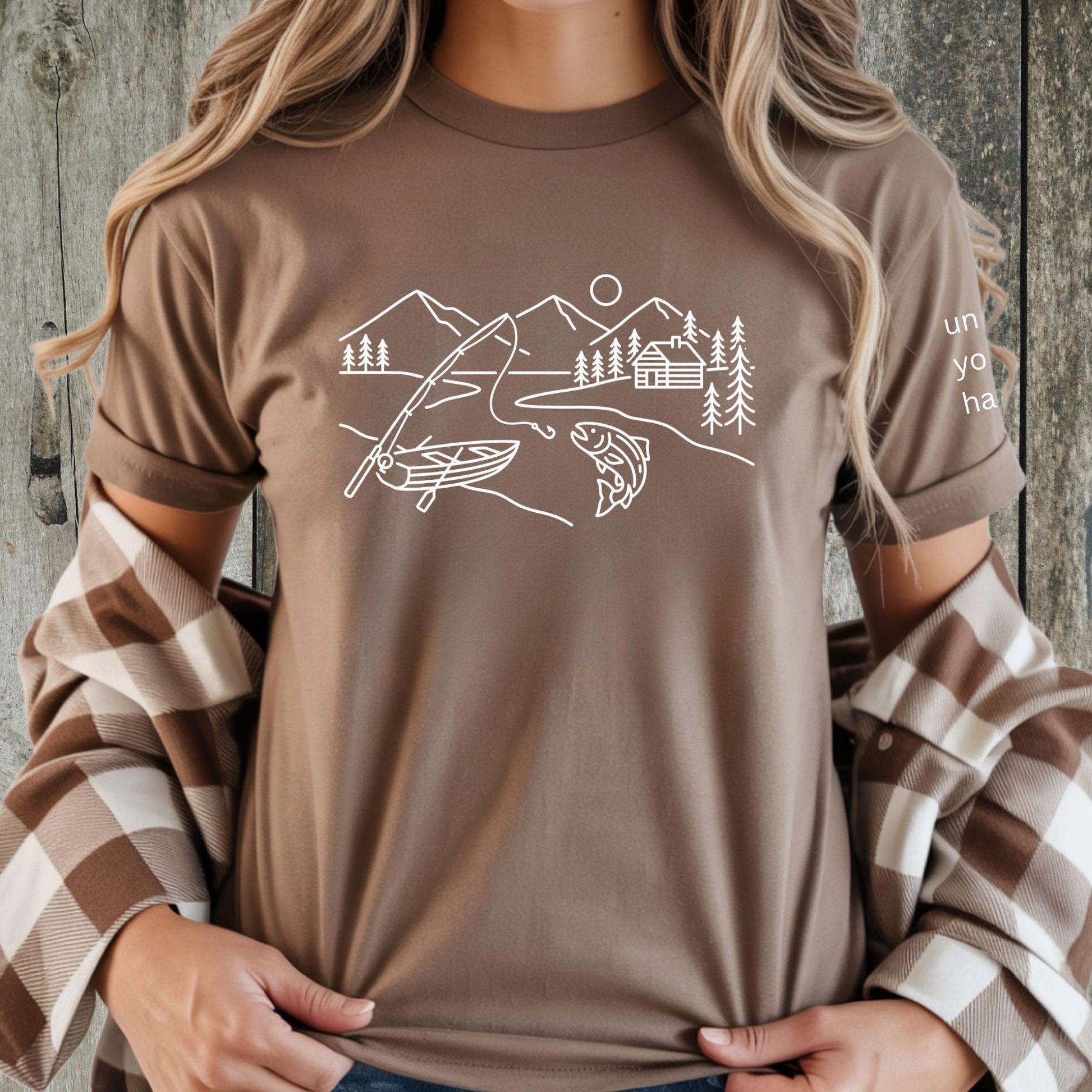 Female wearing brown t-shirt featuring a minimalist white line art fishing design with mountains, river, canoe, fish, cabin, and pine trees.