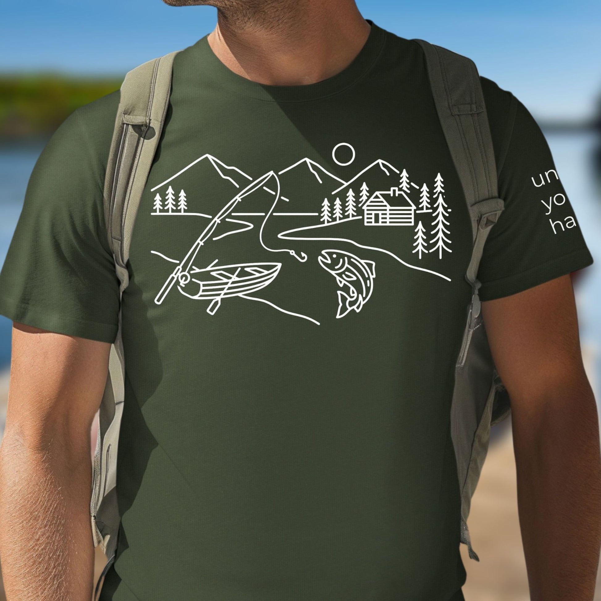 Green t-shirt featuring a minimalist white line art fishing design with mountains, river, canoe, fish, cabin, and pine trees.