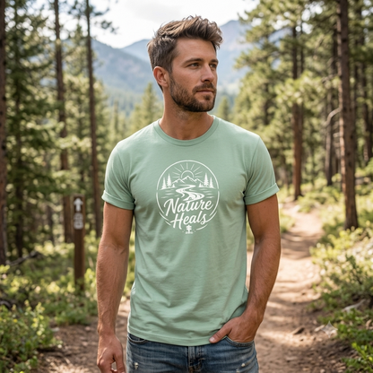 Nature Heals Text with Mountain, Pine Trees, River and Sun Graphic on a sage-green t-shirt on a man on a wooded path