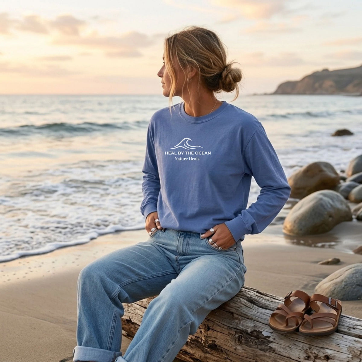 A blue long-sleeve t-shirt with a white graphic on the chest showing an ocean wave and the text 'I HEAL BY THE OCEAN  Nature Heals' on a female by the ocean.
