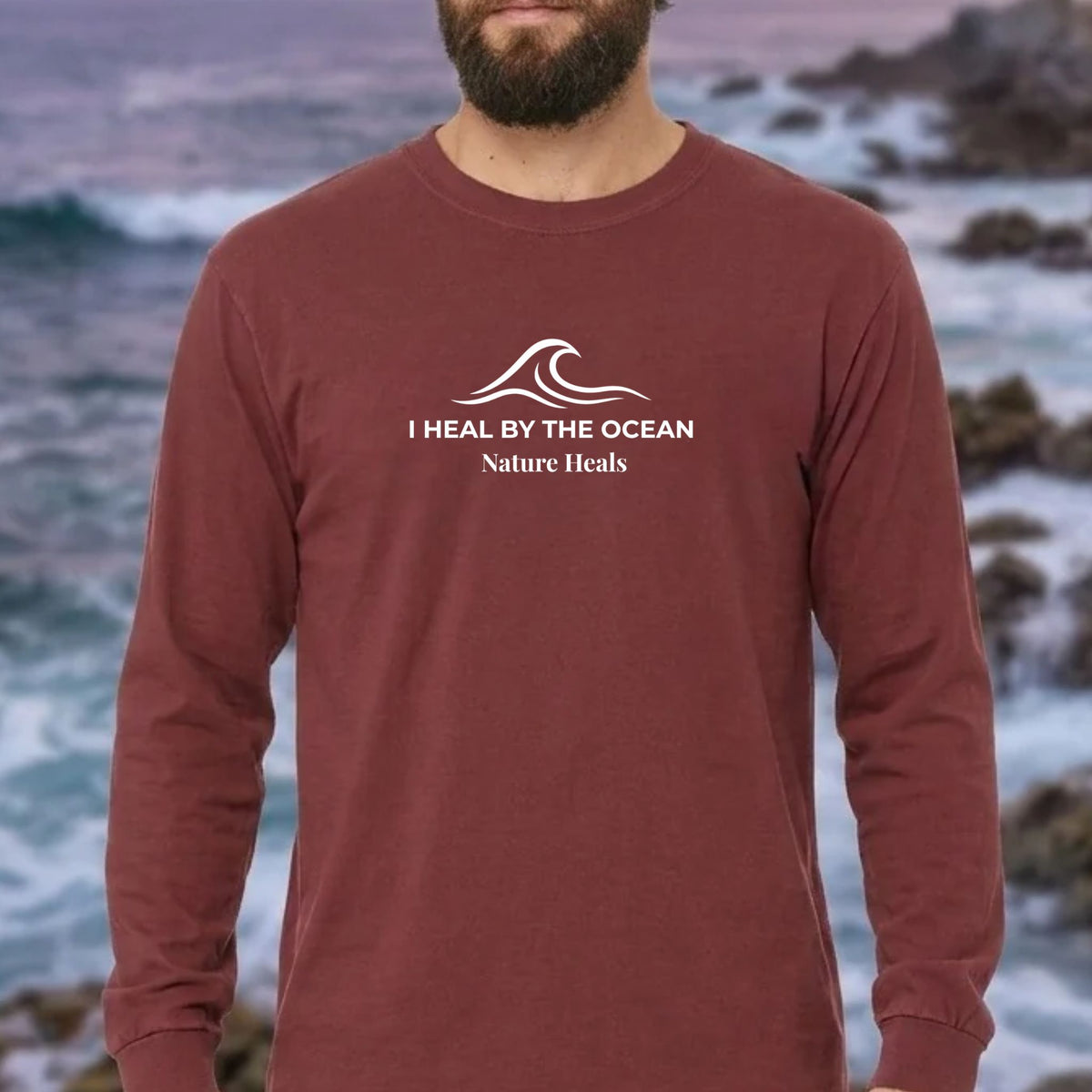 A brick red colored long-sleeve t-shirt with a white graphic on the chest showing an ocean wave and the text 'I HEAL BY THE OCEAN  Nature Heals' on a male by the ocean.