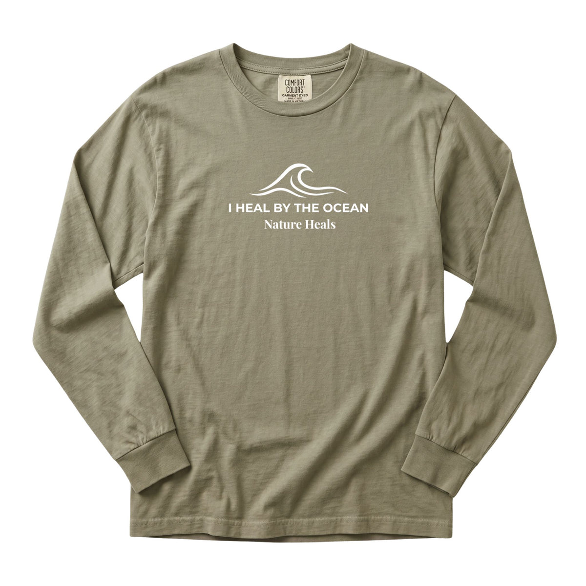 A sandstone colored long-sleeve t-shirt with a white graphic on the chest showing an ocean wave and the text 'I HEAL BY THE OCEAN  Nature Heals'.