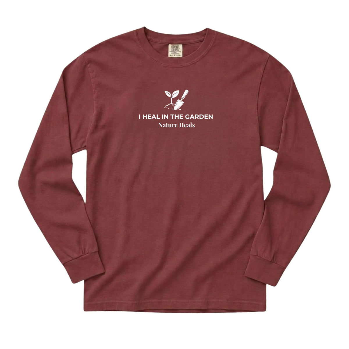 A brick red colored long-sleeve t-shirt with a white graphic on the chest showing a mound of soil, plant and trowel  and the text 'I HEAL IN THE GARDEN  Nature Heals'.