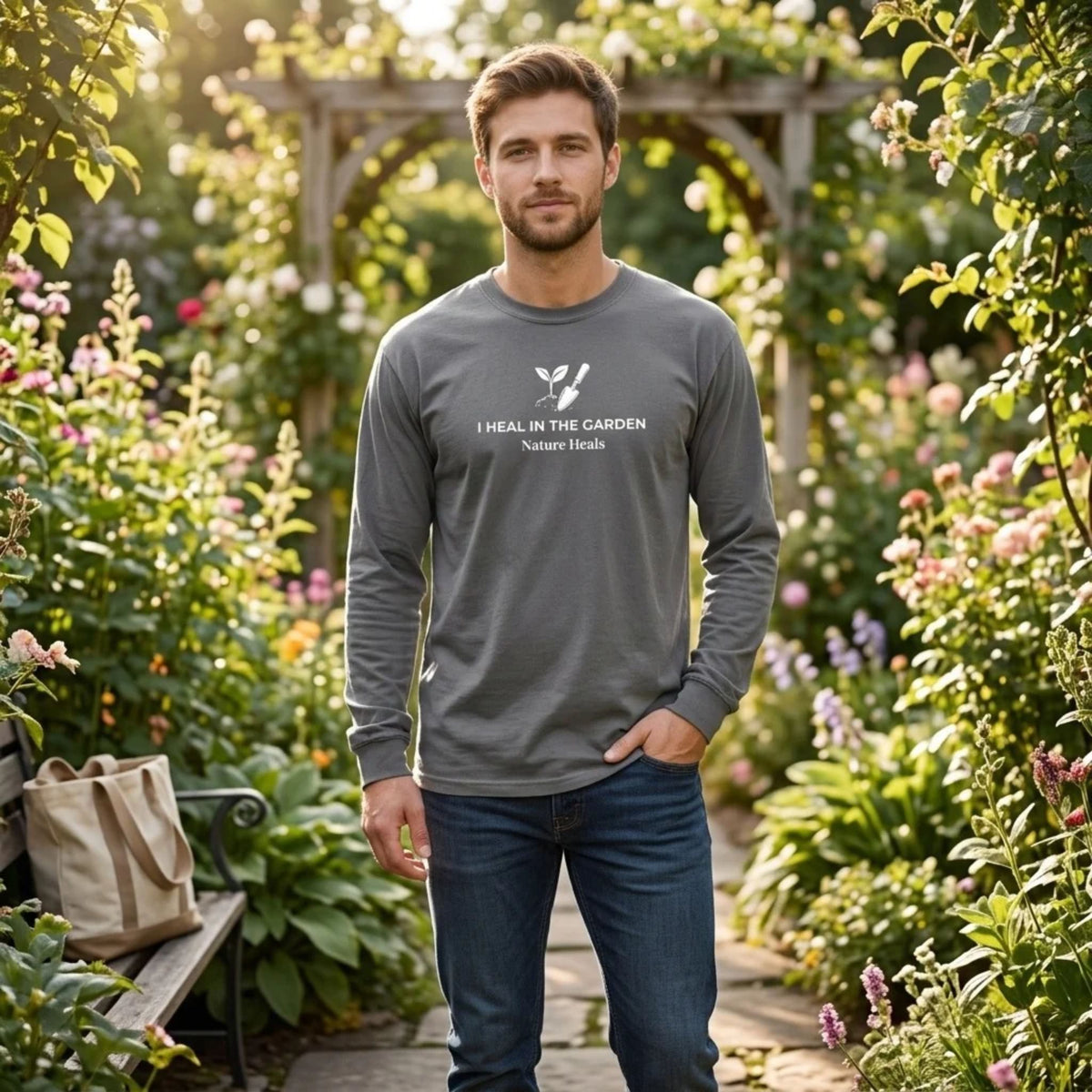 A gray long-sleeve t-shirt with a white graphic on the chest showing a mound of soil, plant and trowel  and the text 'I HEAL IN THE GARDEN  Nature Heals' on a male standing in a garden.