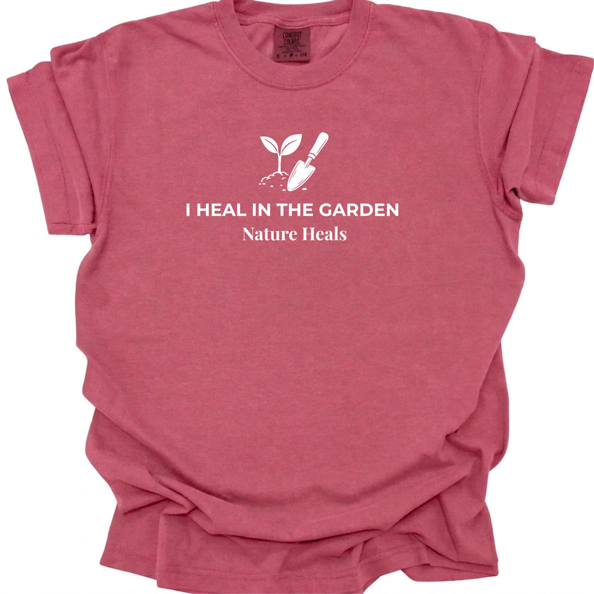 A brick red short-sleeve t-shirt with a white graphic print on the chest featuring a small sprout and trowel, with the text 'I HEAL IN THE GARDEN' and 'Nature Heals'.