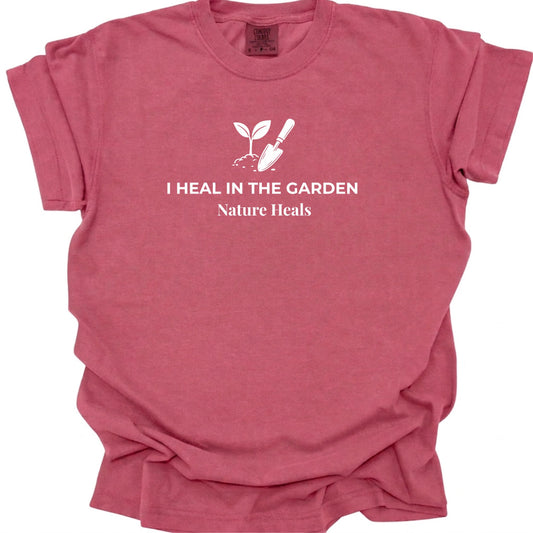 A brick red short-sleeve t-shirt with a white graphic print on the chest featuring a small sprout and trowel, with the text 'I HEAL IN THE GARDEN' and 'Nature Heals'.