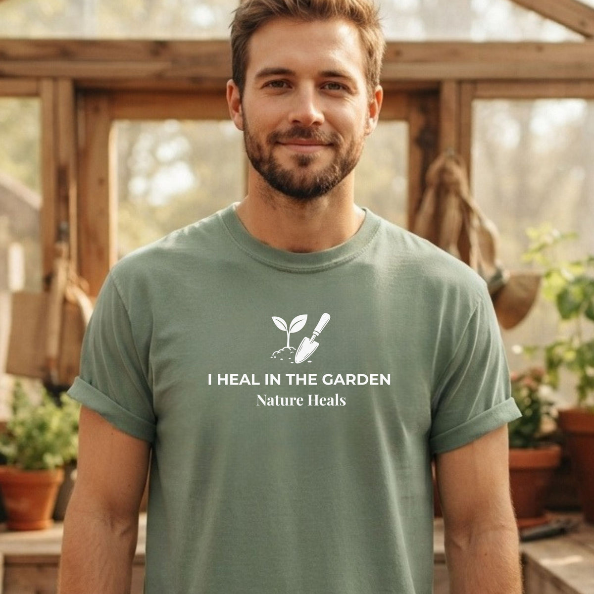 A man wearing a green t-shirt with a white graphic that reads 'I HEAL IN THE GARDEN Nature Heals' and a small sprout and trowel illustration. The background is a warm, softly blurred greenhouse or garden setting.