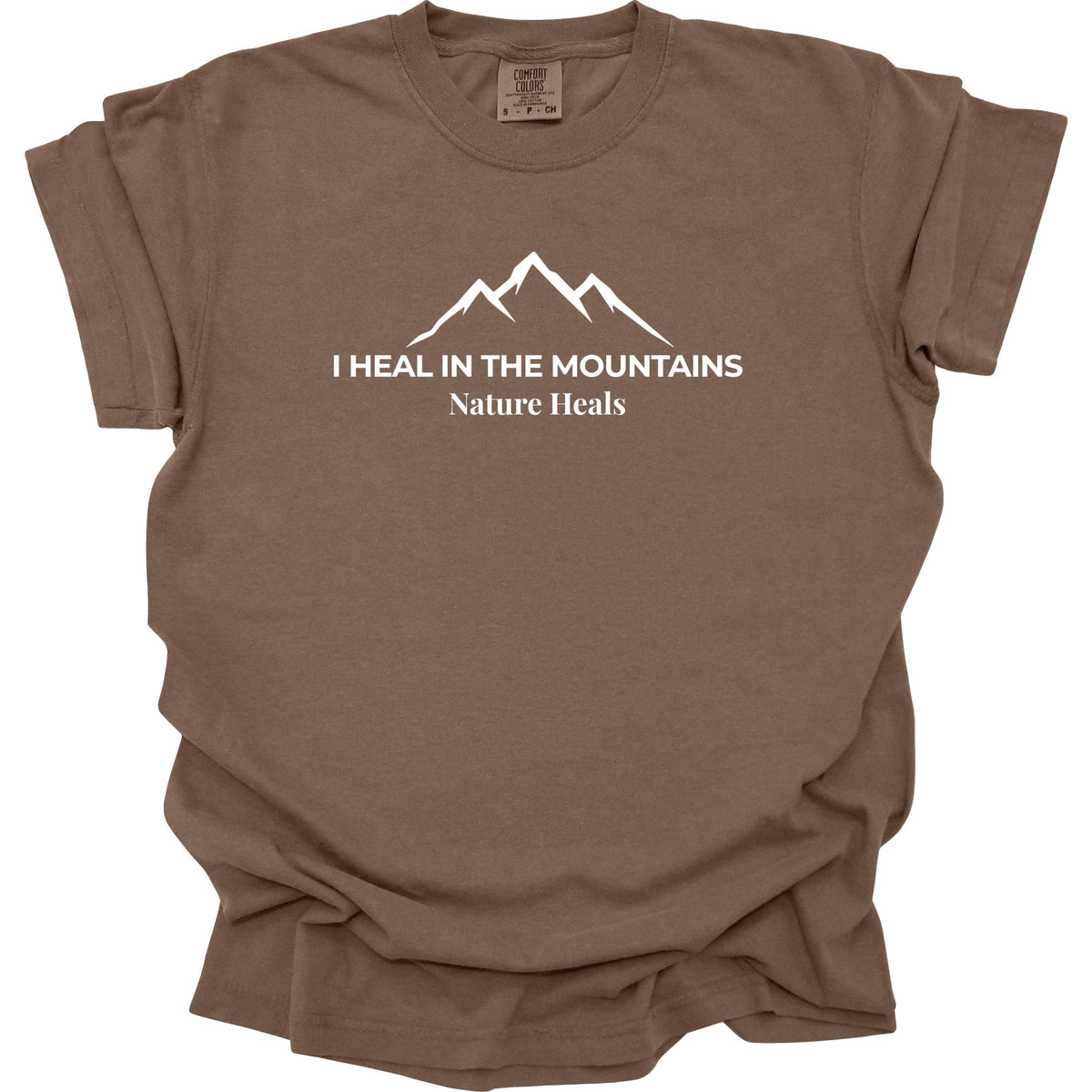 A brown colored short-sleeve t-shirt with a white graphic of a mountain range and the text 'I HEAL IN THE MOUNTAINS Nature Heals' printed on the chest, displayed against a solid white background.
