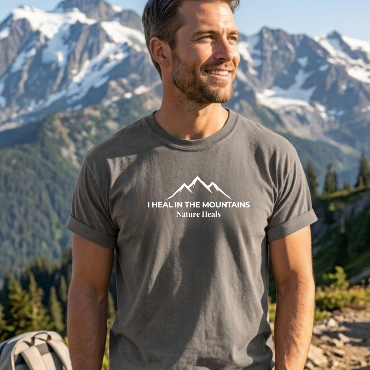 A man wearing a  gray t-shirt with a white mountain graphic and the text 'I HEAL IN THE MOUNTAINS Nature Heals' stands outdoors in front of a scenic mountain background.