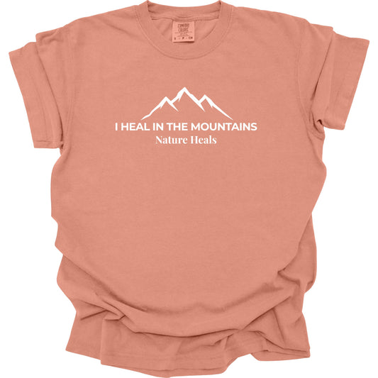 A terracotta-colored short-sleeve t-shirt with a white graphic of a mountain range and the text 'I HEAL IN THE MOUNTAINS Nature Heals' printed on the chest, displayed against a solid white background.