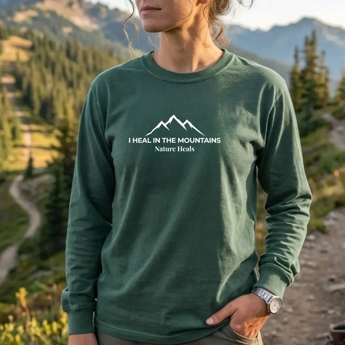 A female wearing a blue spruce green shirt with a mountain graphic and the text 'I HEAL IN THE MOUNTAINS Nature Trails' stands in front of a beautiful mountain landscape with a lake and hiking gear.