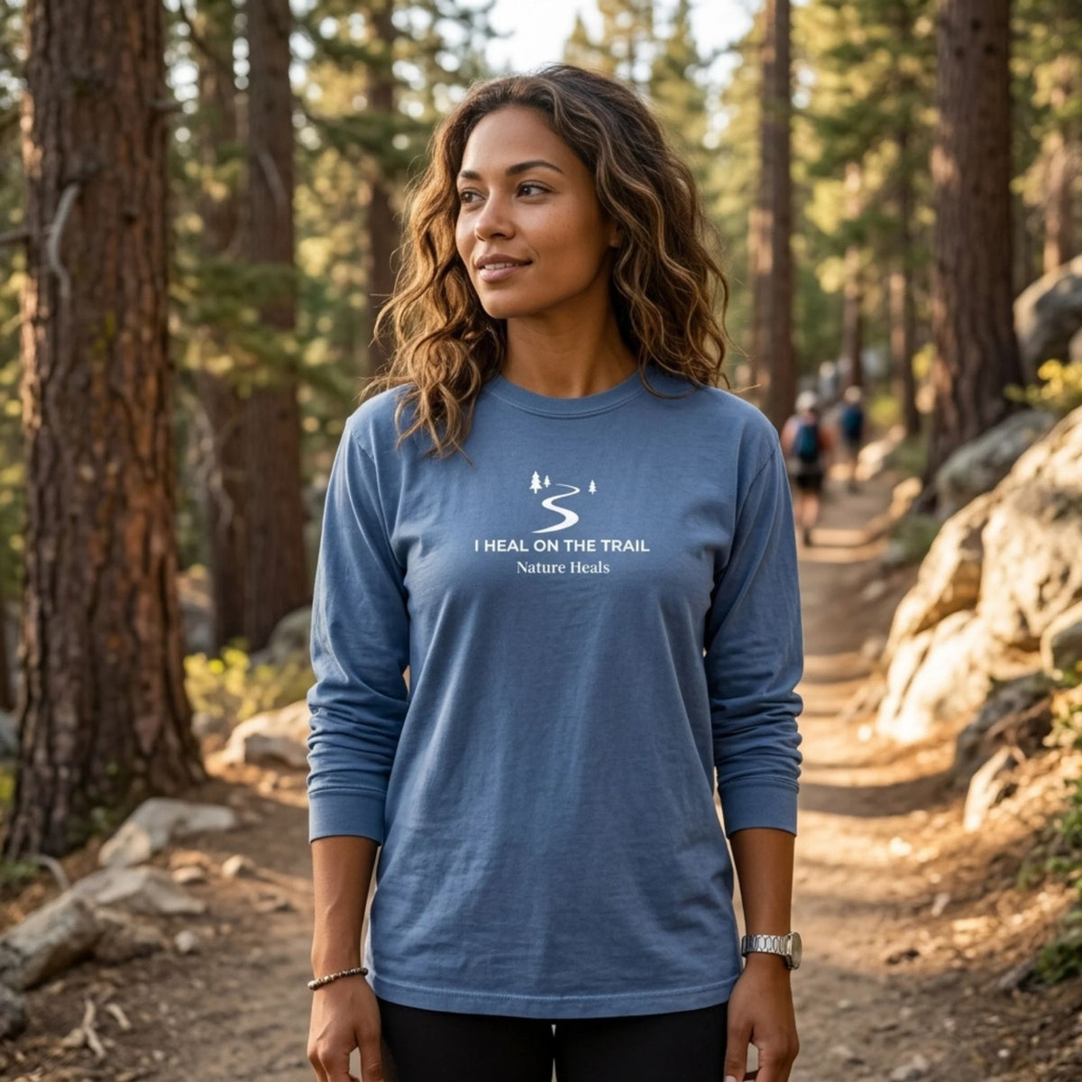 A woman stands on a forest hiking trail wearing a blue long-sleeve t-shirt with a white graphic of a trail and trees that reads 'I HEAL ON THE TRAIL Nature Heals'.