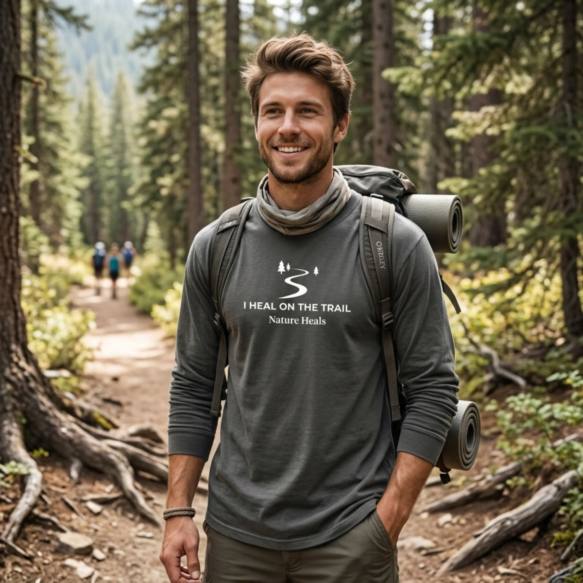 A man stands on a forest hiking trail wearing a dark gray long-sleeve t-shirt with a white graphic of a trail and trees that reads 'I HEAL ON THE TRAIL Nature Heals'.