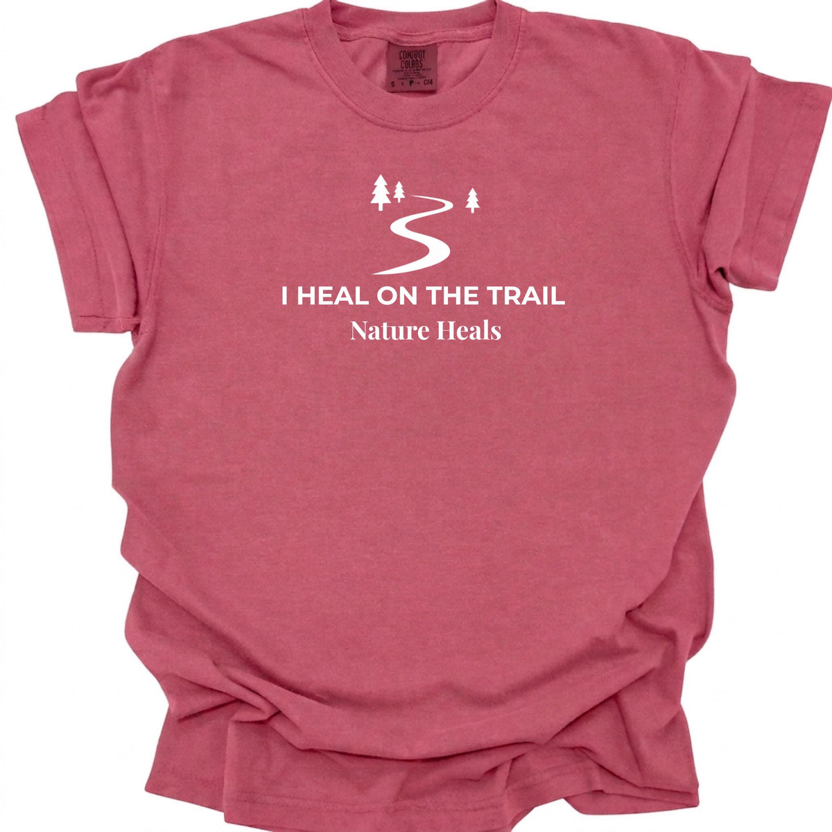 A brick red t-shirt featuring white text that reads 'I HEAL ON THE TRAIL Nature Heals' and a trail icon graphic, with a white background.