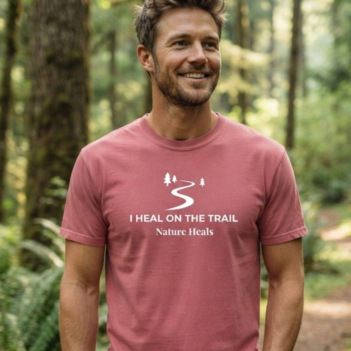 A smiling man wearing a brick red t-shirt with a white graphic that reads 'I HEAL ON THE TRAIL Nature Heals' and a trail icon, standing in a blurred forest setting.