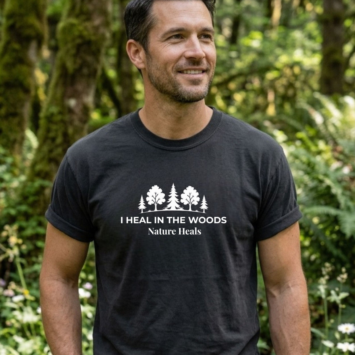 A man wearing a black crewneck t-shirt with a white graphic print that reads 'I HEAL IN THE WOODS Nature Heals' with a forest tree design, set against a blurred green forest background.