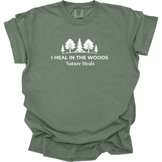 An moss green crewneck t-shirt with a white graphic of trees and the text 'I HEAL IN THE WOODS Nature Heals' printed on the chest, displayed flat against a white background.