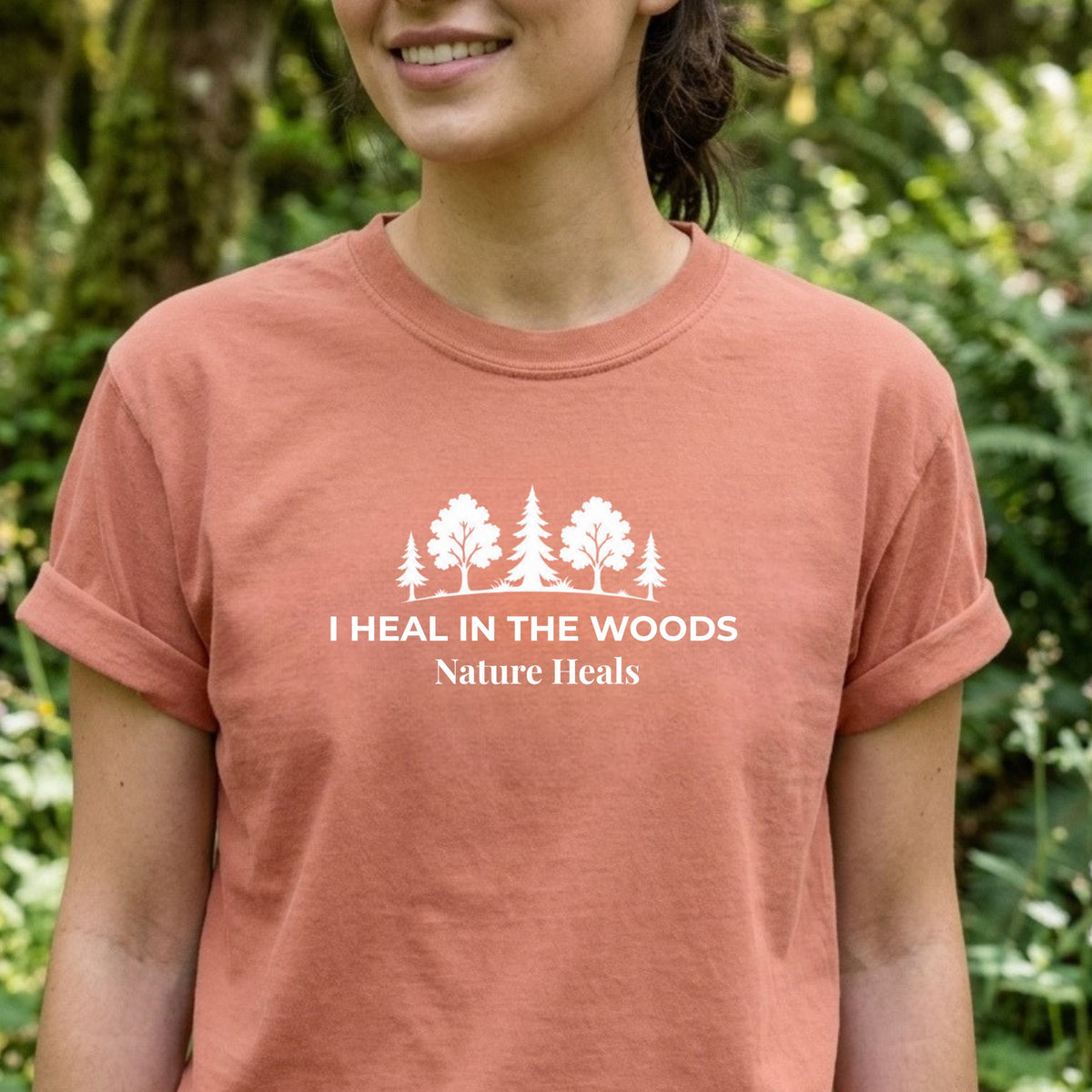 A woman wearing a terracotta-colored t-shirt with a white graphic of trees and the text 'I HEAL IN THE WOODS Nature Heals', set against a blurred green forest background.
