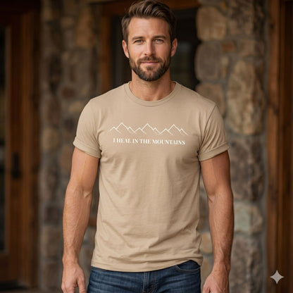 Man wearing a tan t-shirt with 'I Heal  in the Mountains' text and mountain design, standing in front of a stone wall.