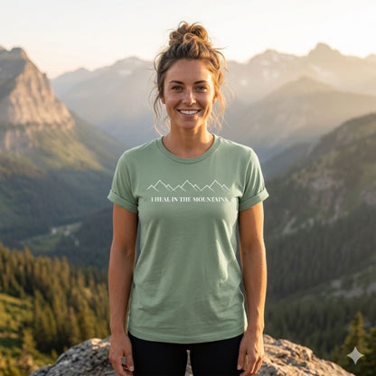 Woman wearing a green t-shirt with mountain design and text "I HEAL IN THE MOUNTAINS", standing in front of a mountainous landscape.