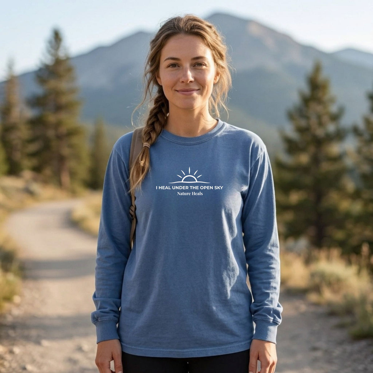 A woman standing outdoors on a dirt path with mountains and trees in the background, wearing a blue long-sleeve shirt with a white sunrise graphic and the text 'I HEAL UNDER THE OPEN SKY Nature Health' on the chest.