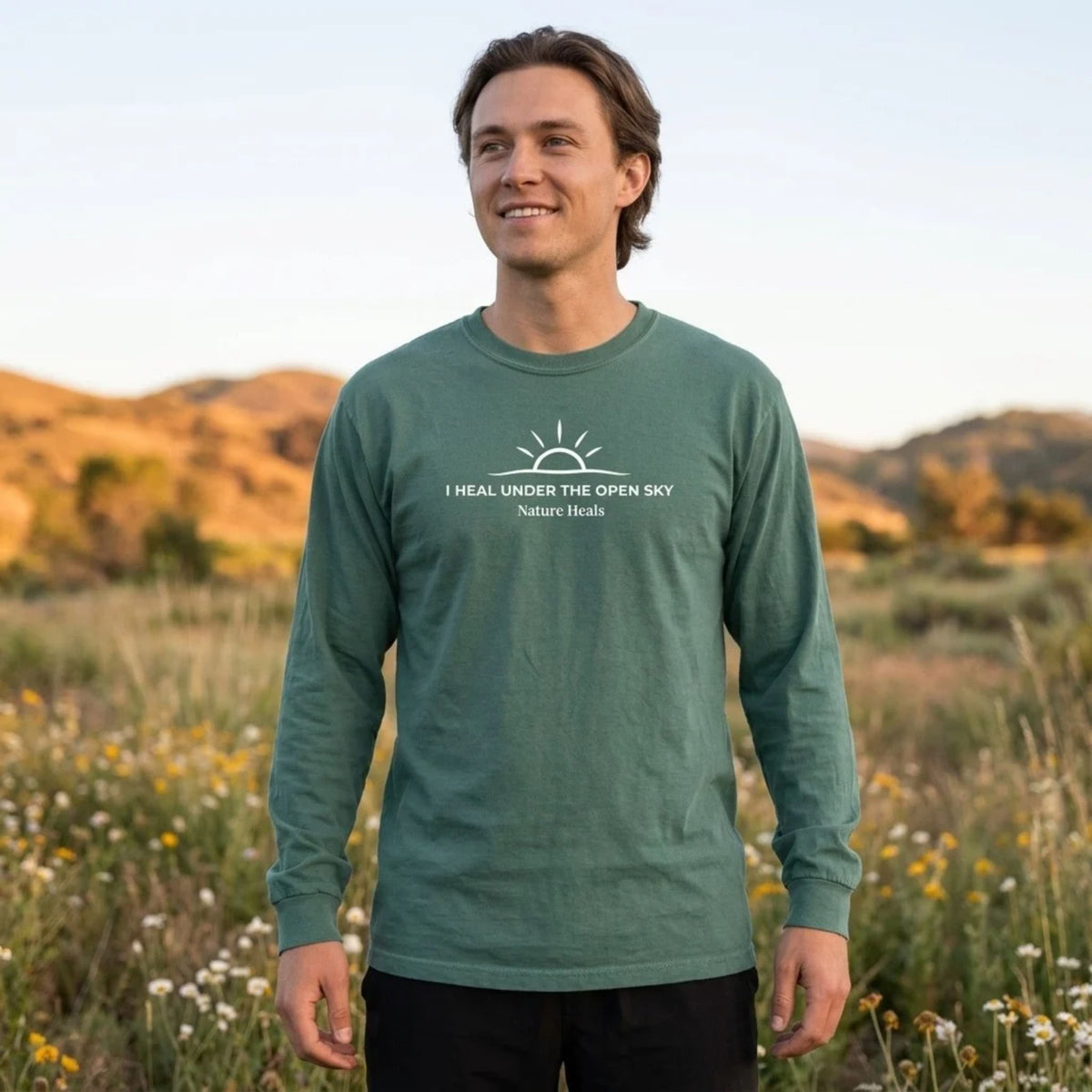 A man wearing a sage green long-sleeve t-shirt with a white graphic that reads 'I HEAL UNDER THE OPEN SKY Nature Heals' standing in an outdoor grassy field at sunset.