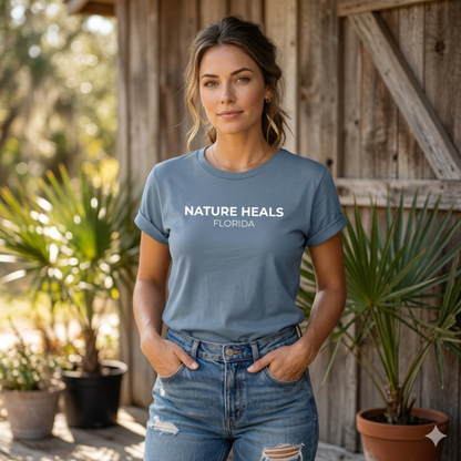 Woman wearing a blue t-shirt with 'NATURE HEALS FLORIDA' text, standing in front of a wooden building and plants.