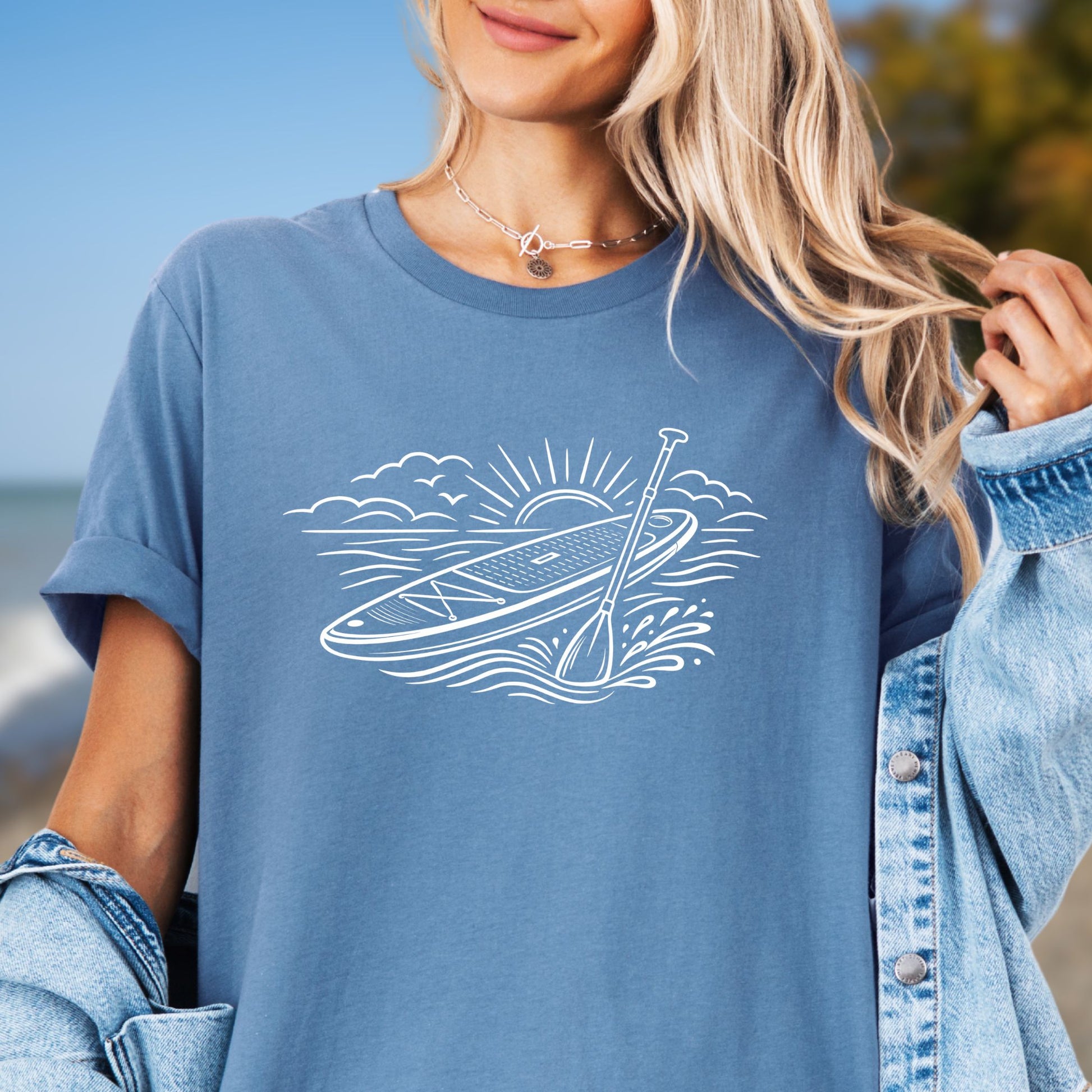 Blue t-shirt featuring white line art of a stand up paddle board with paddle on water at sunrise, worn by model, ocean and beach lifestyle design