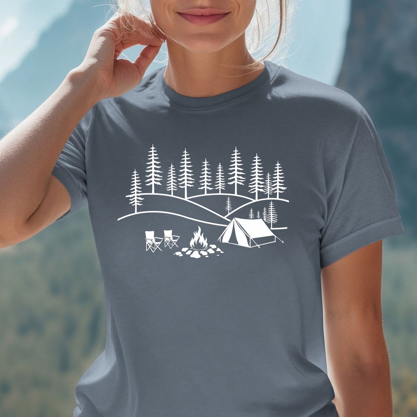 Minimalist camping graphic t-shirt with forest trees, tent, campfire and chairs design on blue shirt against a blurred background.