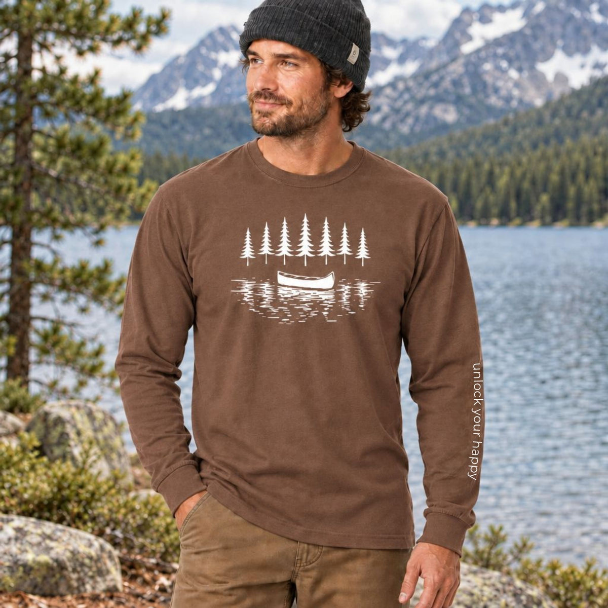 A man wearing a brown long-sleeve t-shirt with a white graphic of pine trees and a canoe on the chest, with 'unlock your happy' text on the sleeve, standing outdoors in front of a mountain lake.