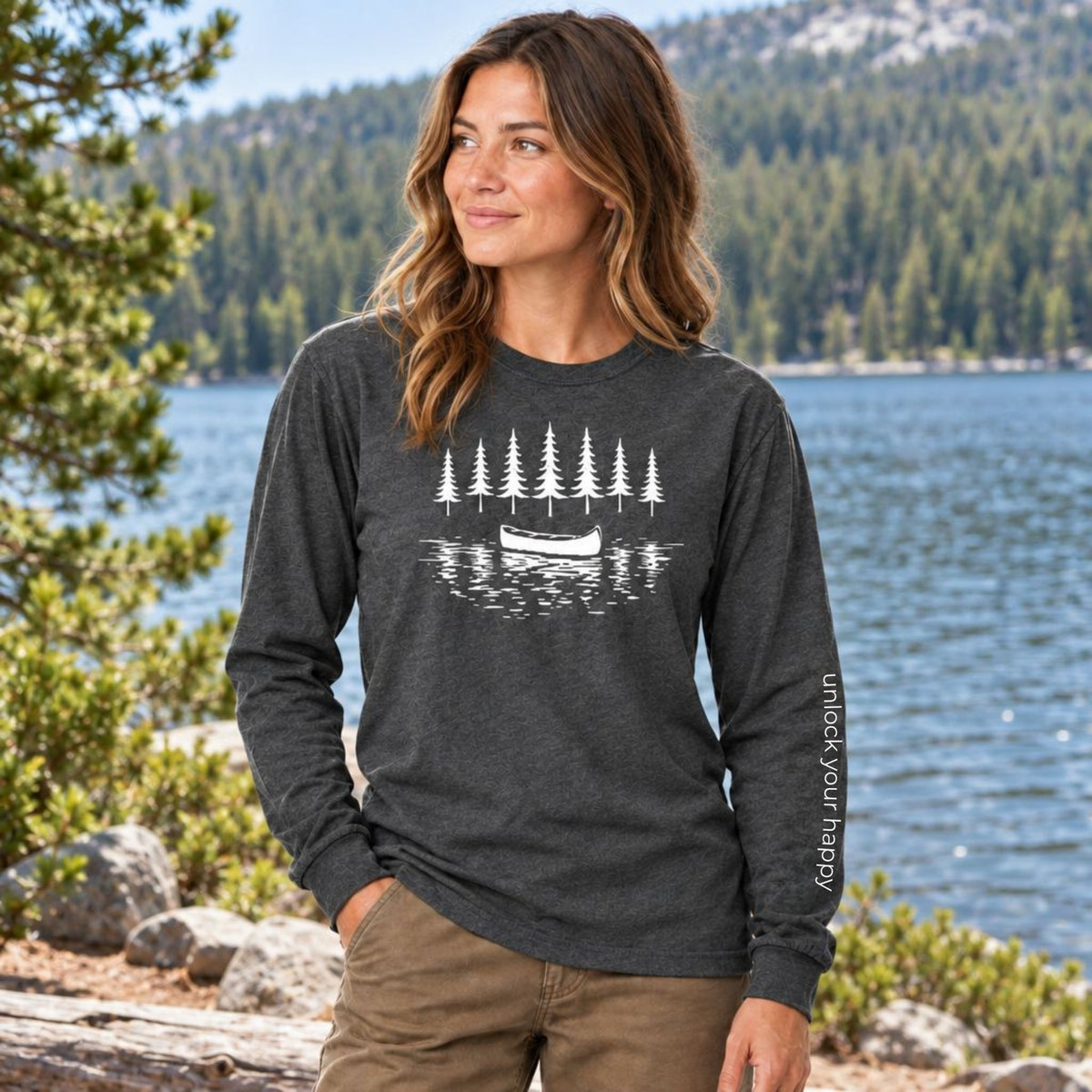 A woman standing outdoors by a lake with trees and mountains in the background, wearing a pepper grey  long-sleeve shirt with a white graphic of pine trees and a canoe, with 'unlock your happy' text on sleeve. 