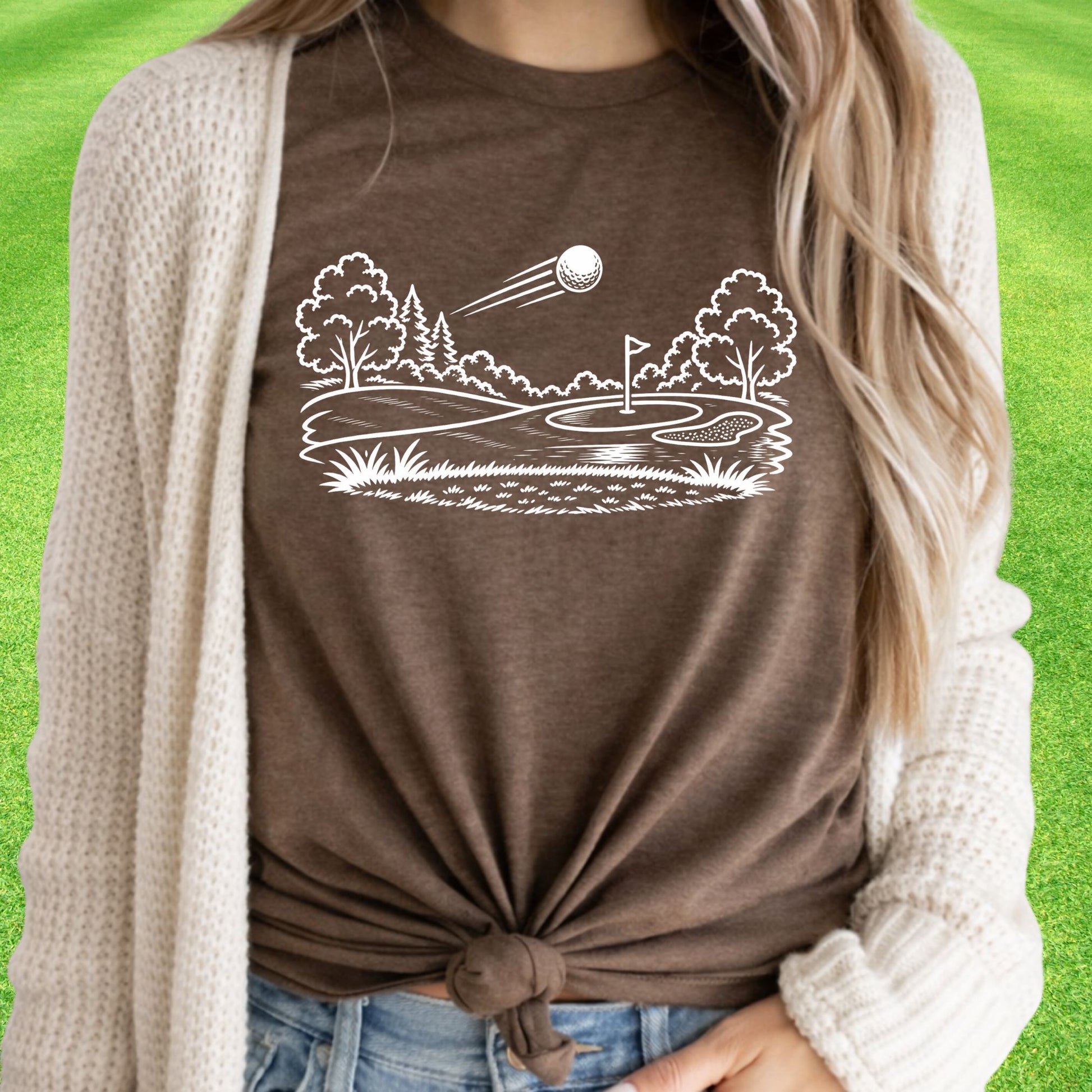 Minimalist golf course graphic t-shirt with line art design of golf ball and green on soft unisex brown tee on green lawn