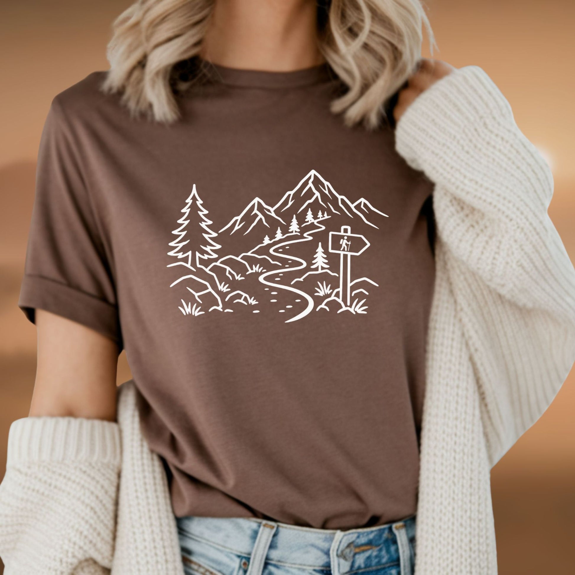 Person wearing a minimalist brown t-shirt with white mountain and forest trail design, hiking path graphic for nature lovers and outdoor enthusiasts