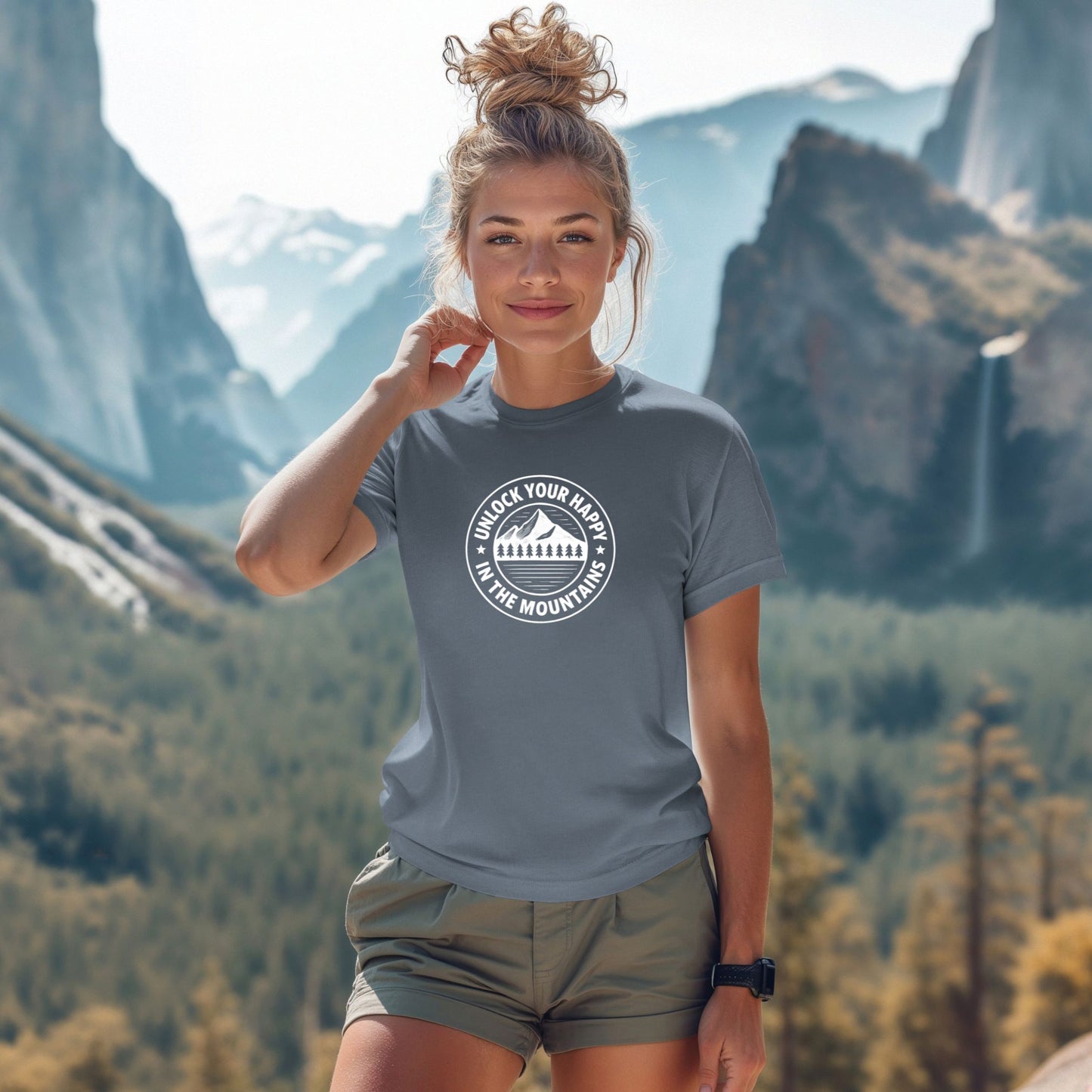 Blue unisex graphic t-shirt featuring a circular mountain and pine tree design with the text “Unlock Your Happy in the Mountains” on a female in the mountains.