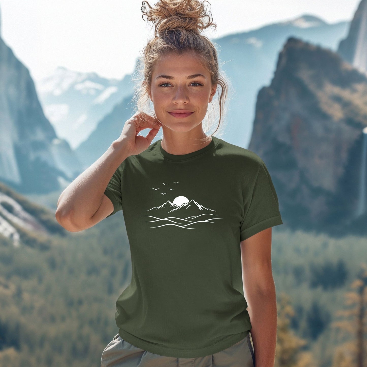 Woman wearing a military green mountain graphic t-shirt with minimalist white line art design, outdoors with mountains in the background
