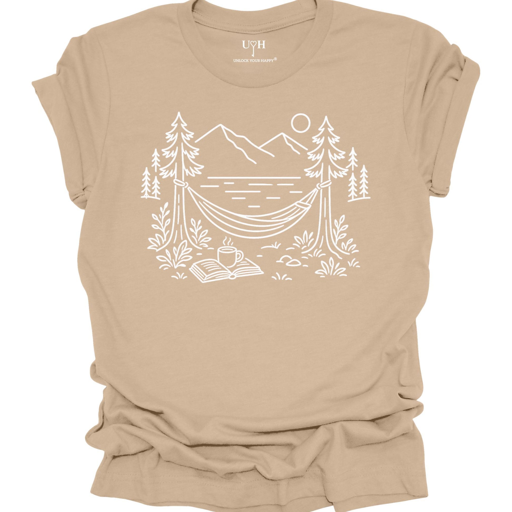 Tan unisex t-shirt with a white line art hammock scene featuring mountains, trees, a lake, coffee cup, and open book, plus ‘unlock your happy’ on the sleeve.