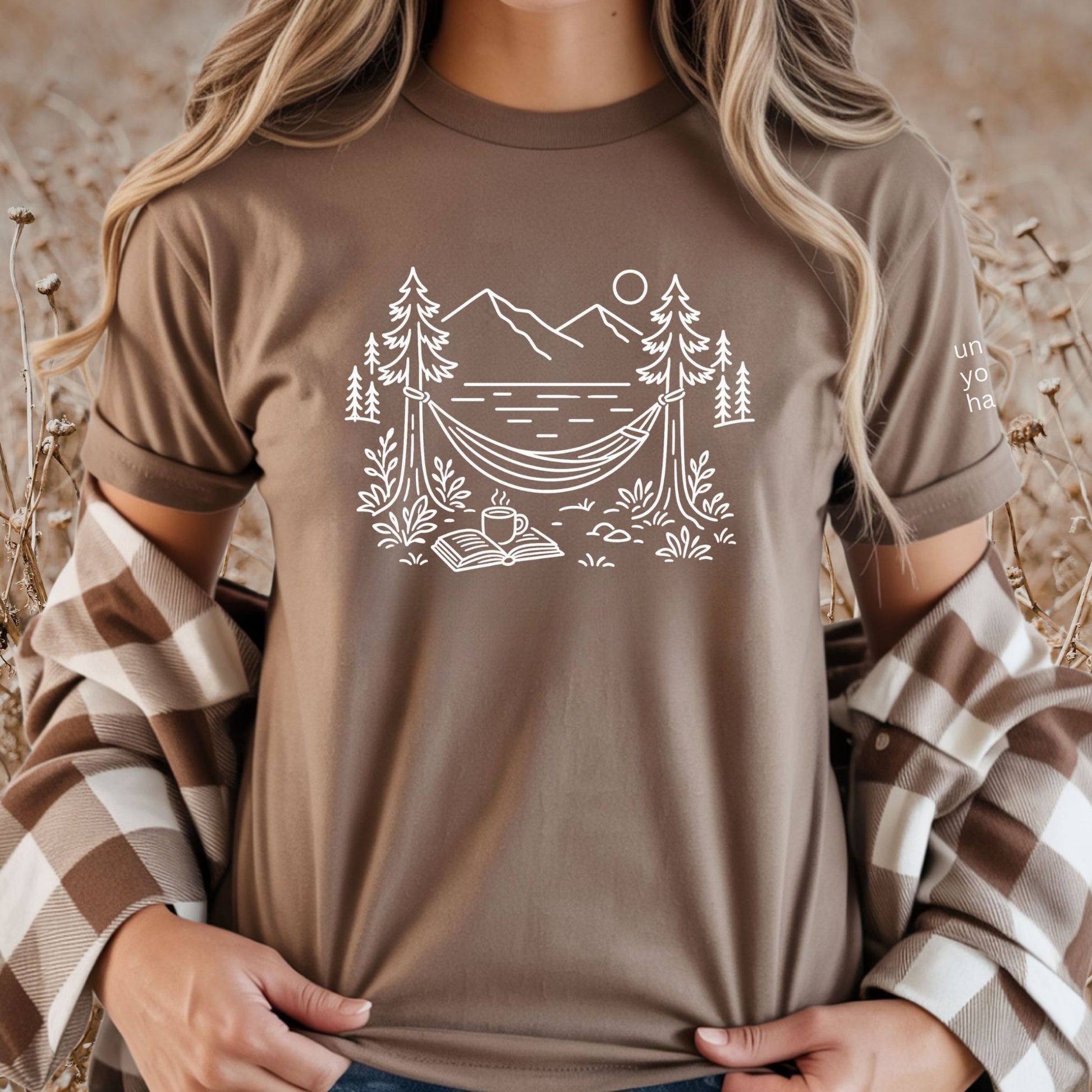 Brown unisex t-shirt with a white line art hammock scene featuring mountains, trees, a lake, coffee cup, and open book, plus ‘unlock your happy’ on the sleeve on a female.