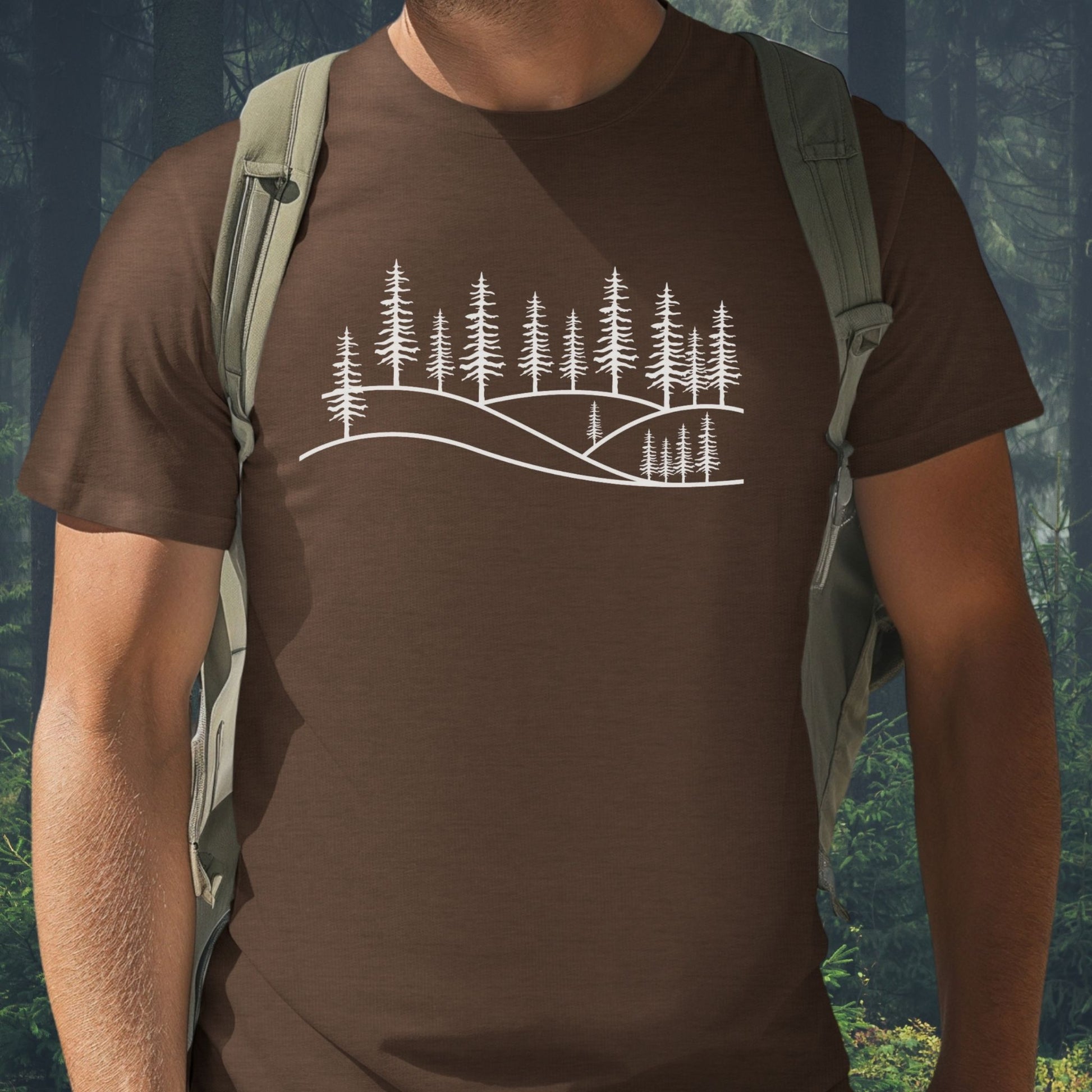A man wearing a chocolate brown nature graphic t-shirt with a white pine forest line art design, pictured against a lush green woodland background. Minimalist apparel for mental well-being and hiking.