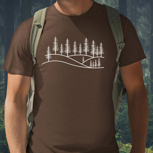 A man wearing a chocolate brown nature graphic t-shirt with a white pine forest line art design, pictured against a lush green woodland background. Minimalist apparel for mental well-being and hiking.