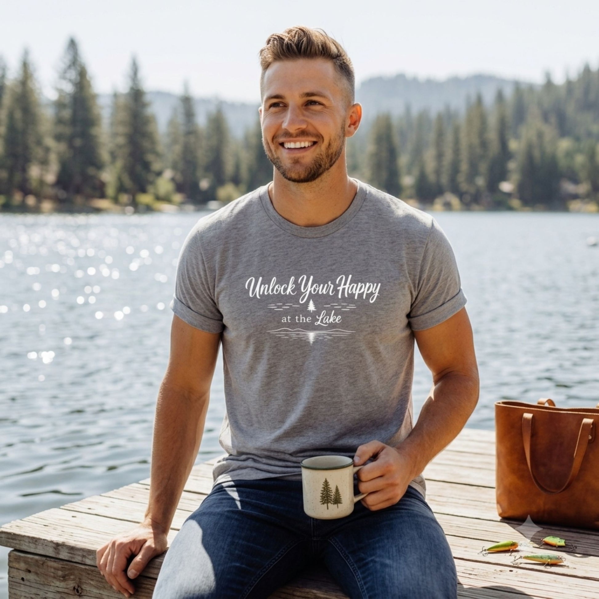 A smiling man sitting on a wooden dock by a lake wears a heather gray t-shirt with the white printed text 'Unlock Your Happy at the Lake'. He is holding a white mug, with a brown leather tote bag on the dock next to him and a forested lake in the background.