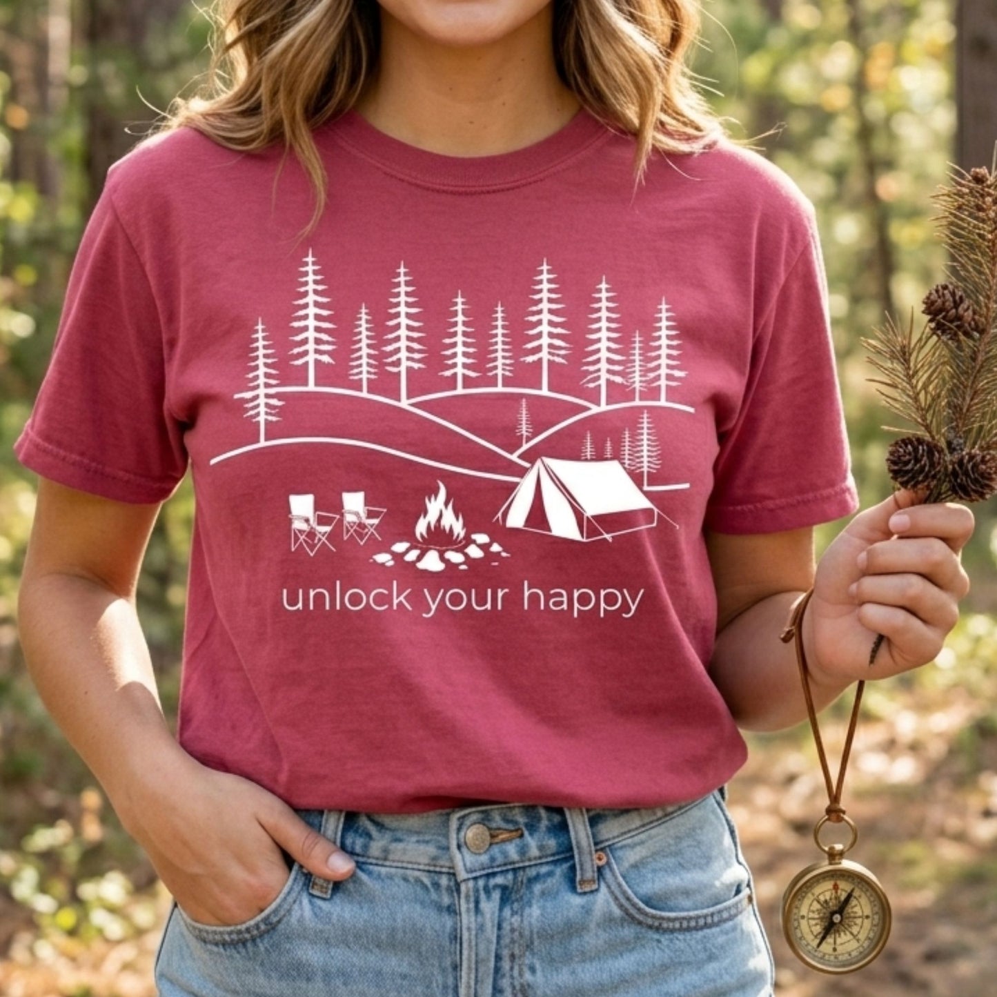 Person wearing a brick red t-shirt with a camping-themed design and unlock your happy text, standing in a forest.