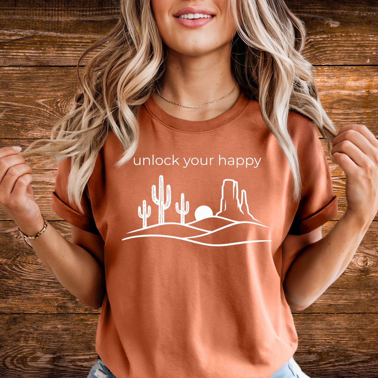 Woman wearing a terracotta-colored t-shirt with a desert landscape graphic and text 'unlock your happy' against a wooden background.