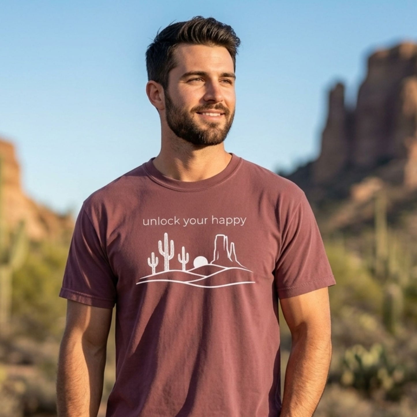Man wearing a brick colored t-shirt with a desert-themed design and text, standing in a desert landscape.