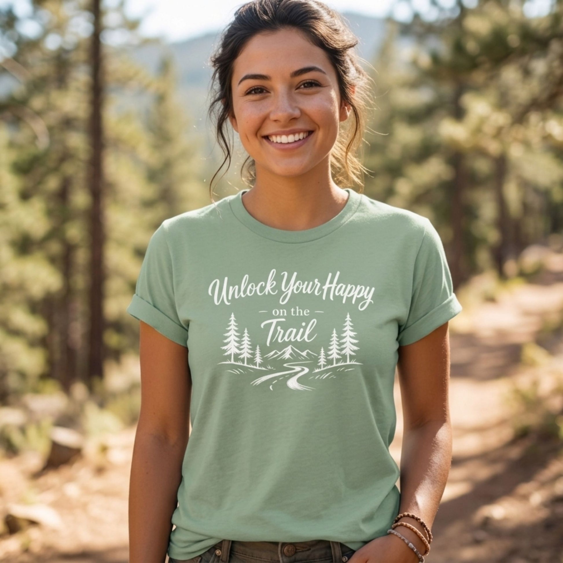 A woman smiling outdoors on a forest trail, wearing a sage green graphic t-shirt with the white text 'Unlock Your Happy on the Trail' and a pine tree illustration.