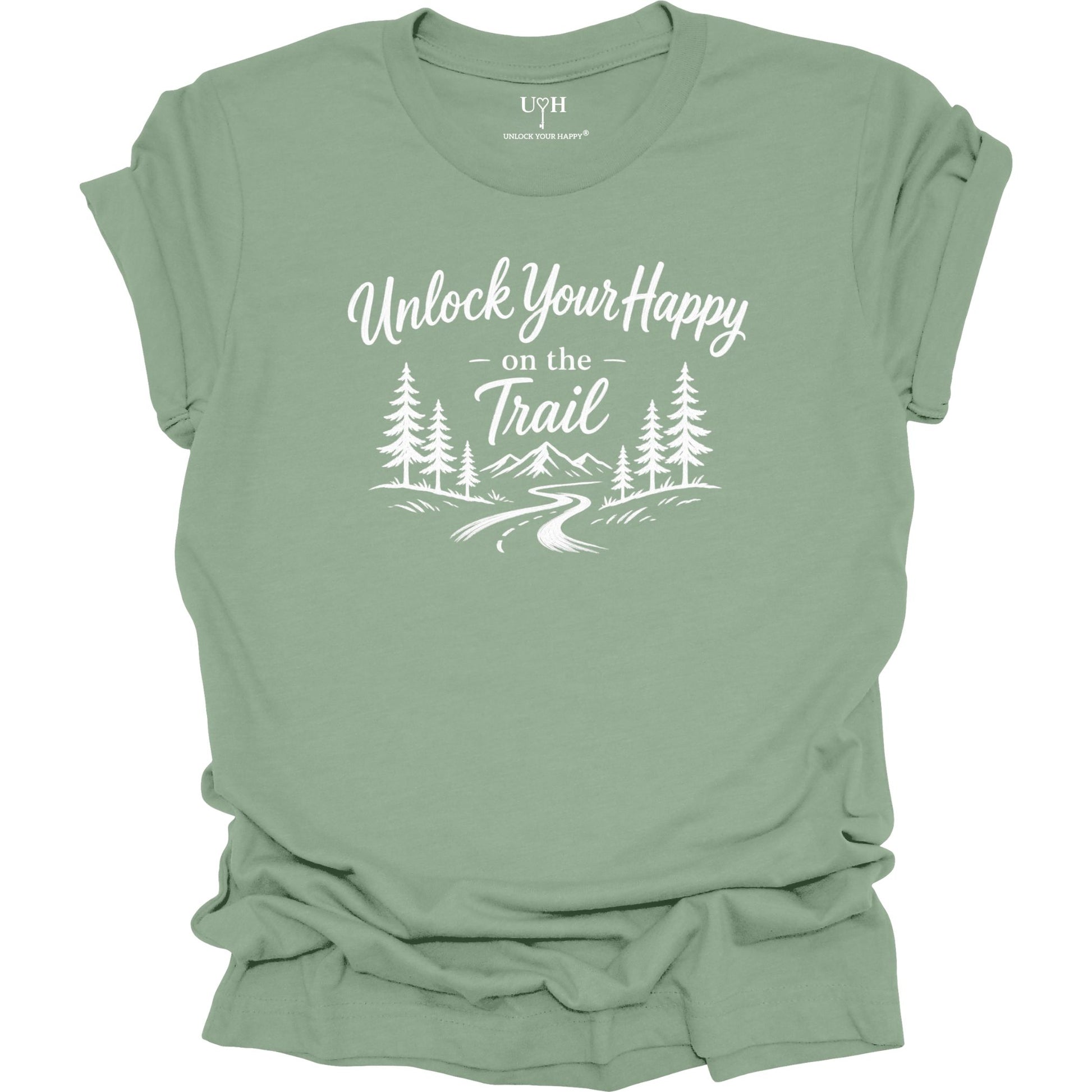 A sage green short-sleeve t-shirt with a white graphic print that reads 'Unlock Your Happy on the Trail' above an illustration of pine trees, mountains, and a winding trail. The shirt is shown flat against a white background, with the sleeves slightly rolled.