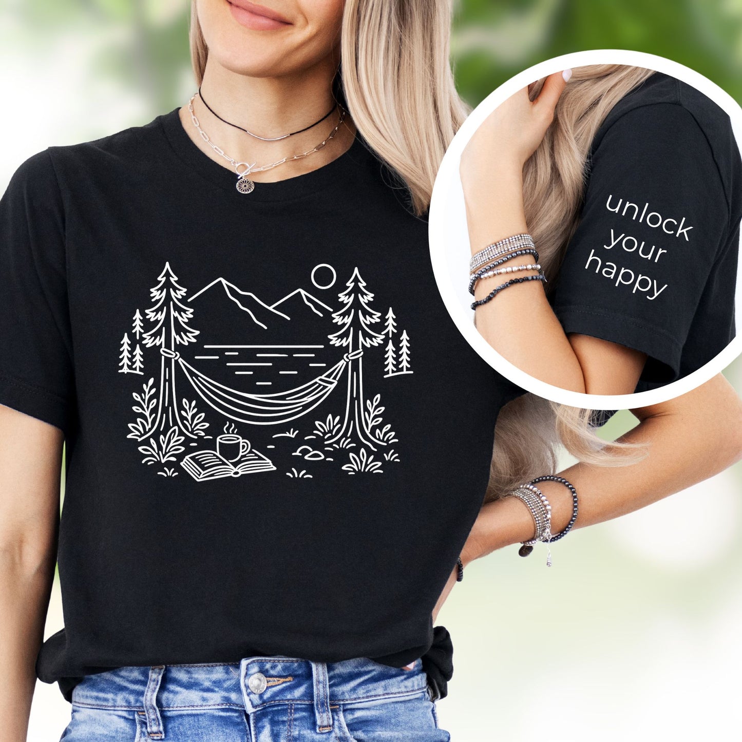 Black unisex t-shirt with a white line art hammock scene featuring mountains, trees, a lake, coffee cup, and open book, plus ‘unlock your happy’ on the sleeve on a female.