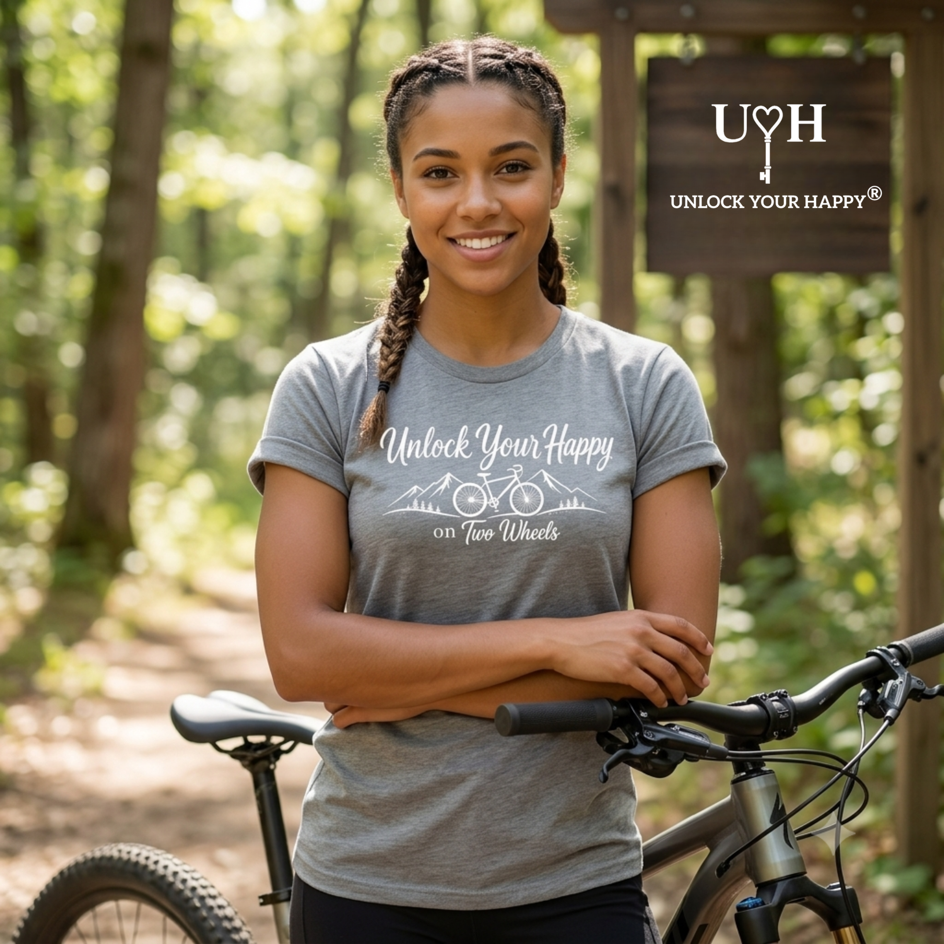 A woman wearing a gray graphic t-shirt stands in a forest with a mountain bike. The t-shirt features the white text 'Unlock Your Happy on Two Wheels' with a bicycle graphic.