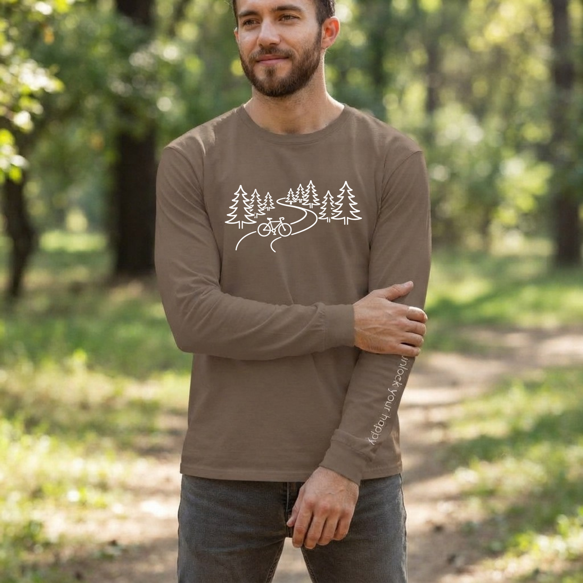 A man wearing a brown long-sleeve t-shirt with a white forest and bicycle graphic on the chest, with "unlock your happy" text on the left sleeve standing in a sunlit forest path.